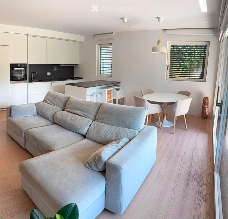 Modern open-plan living and dining area with light-colored furniture, a grey sectional sofa, a white dining table with four chairs, and a sleek kitchen area with white cabinets and a black countertop.