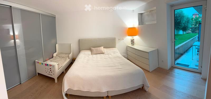 Bright bedroom with two beds, a nightstand with a lamp, and a view of a green area through a large window.