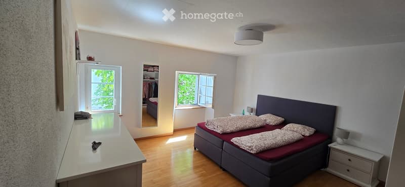 Bright bedroom with wooden flooring, a double bed with pillows and blankets, a nightstand with a lamp, a large window with a view, and a wardrobe with clothes.