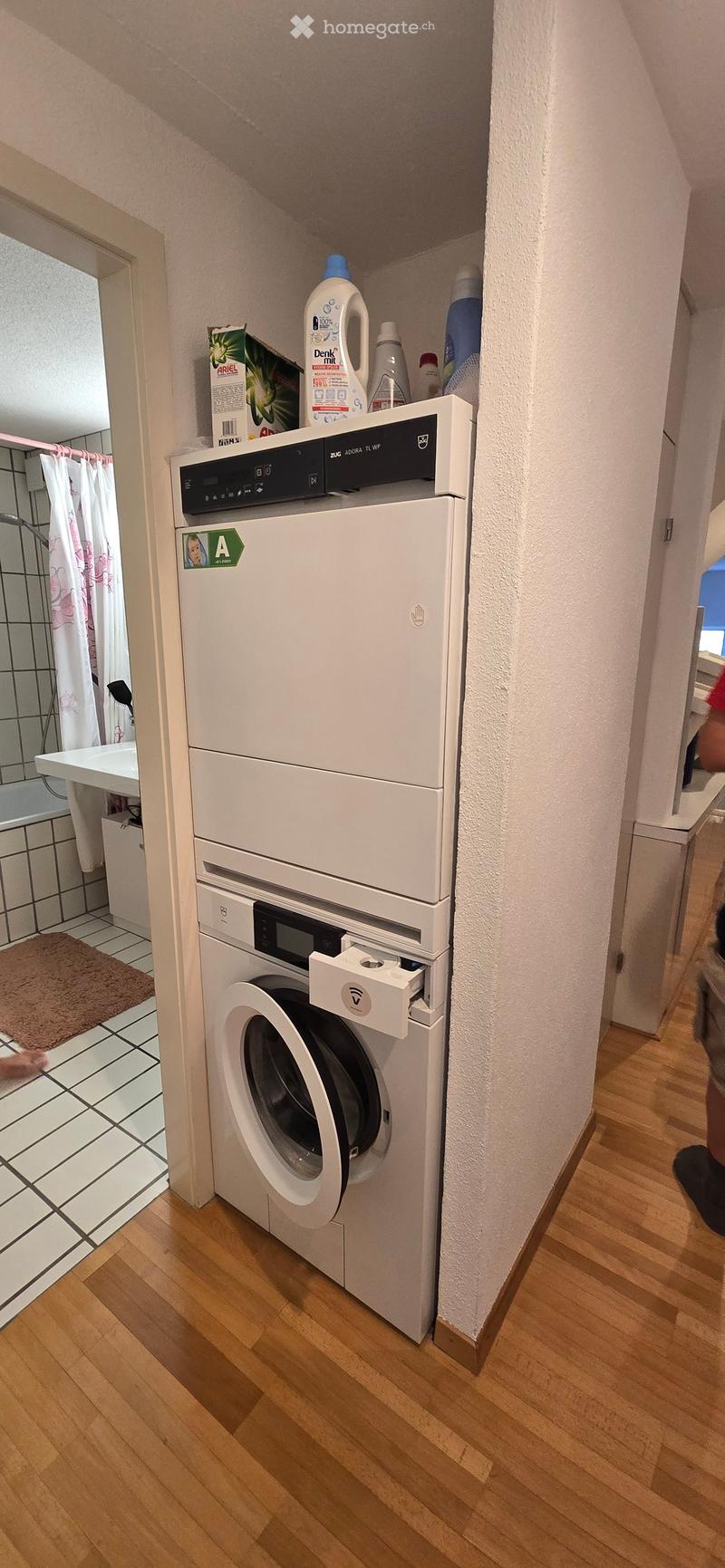 Hallway with a stacked washing machine and dryer unit, wooden flooring, and a small bathroom area visible to the left.