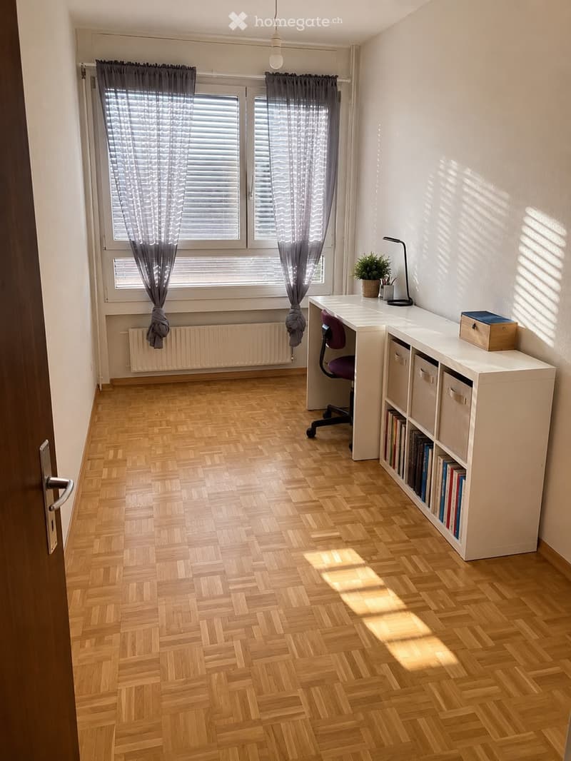 Bright, modern room with wooden herringbone flooring, large windows with grey curtains, a white desk with a chair, a bookshelf filled with books, a small potted plant, and a lamp.