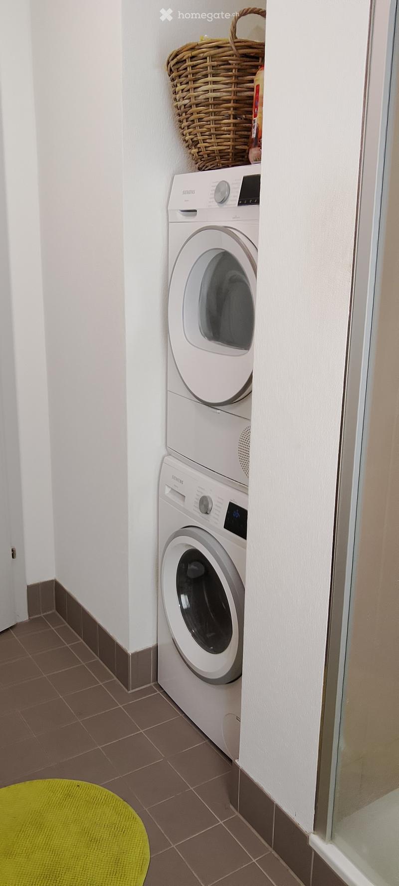 A compact bathroom with a white Siemens washing machine and dryer unit, a wicker basket on top, and a bright green round rug on the floor.