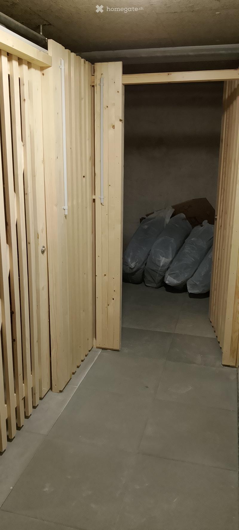 Empty basement space with unfinished wooden paneling and concrete floor, featuring a partially open wooden door leading to a storage area with bags.