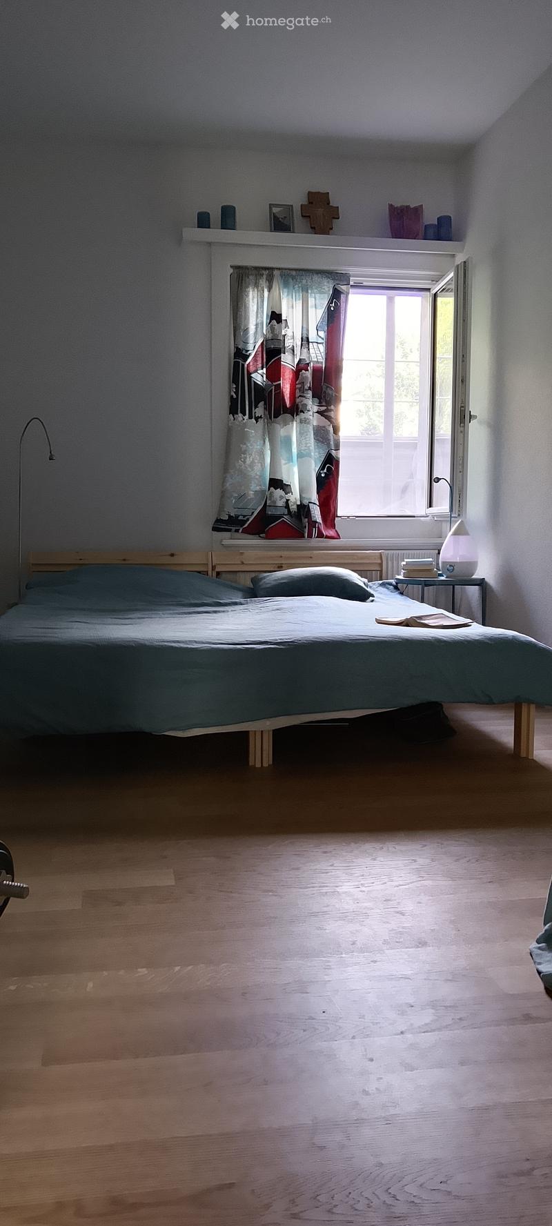 A cozy bedroom with a wooden bed, nightstand, lamp, and a window with a colorful curtain. The room features light wooden flooring and a small shelf above the bed with decorative items.