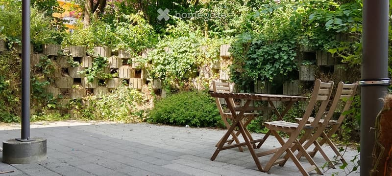 A small outdoor seating area with a wooden table and four folding chairs, surrounded by lush greenery and a stone wall.