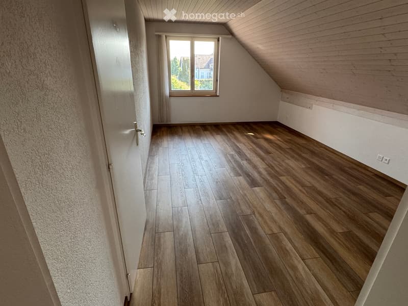 Empty room with wooden flooring, white walls, and a window with curtains. The ceiling has wooden paneling and a sloped design typical of an attic space.