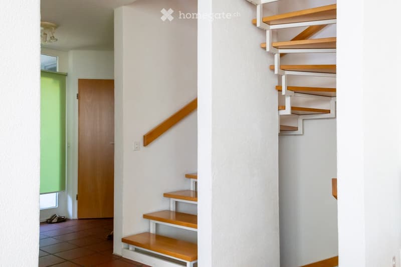 A narrow hallway with a wooden staircase leading upwards, featuring white walls, a wooden handrail, and a tiled floor. A wooden door is visible on the left, and a small window with green blinds is near the entrance.