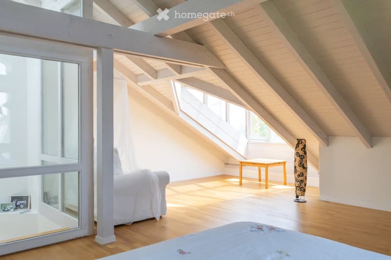 Bright, airy attic room with wooden floors, exposed wooden beams, large windows, a white sofa, a small wooden table, and a decorative vase.
