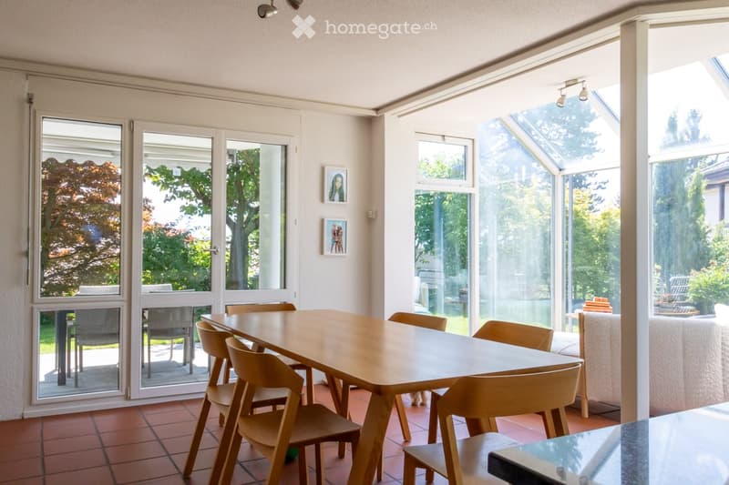 Bright dining area with a large wooden dining table surrounded by chairs, a sofa visible through the glass doors, and large windows offering a view of the garden.