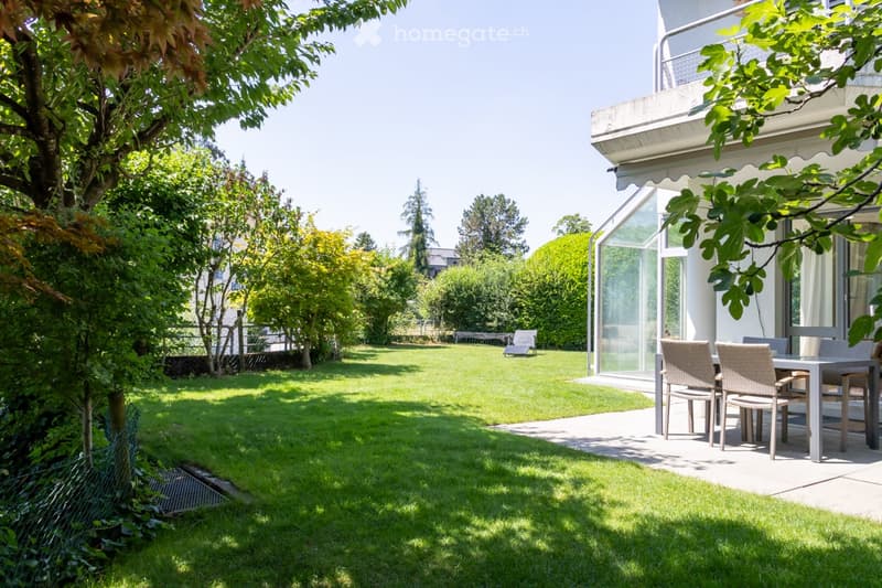 Well-maintained garden with a lawn, trees, and a patio area with outdoor furniture near a house with a balcony.