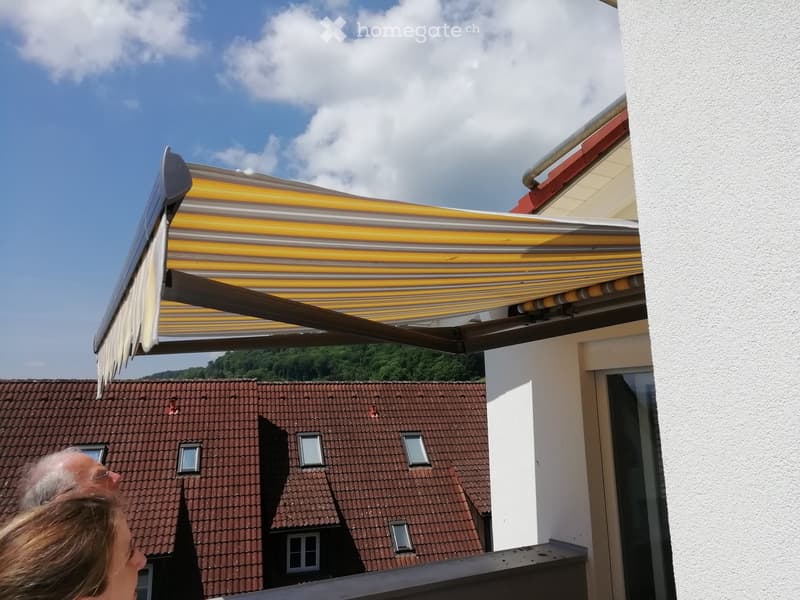 Balcony with a large retractable awning, overlooking a tiled roofed building and a green landscape.
