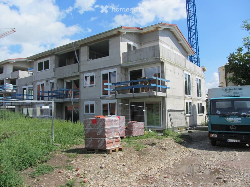 Two-story residential building under construction with exposed concrete walls, large windows, and blue railings on balconies. Stacks of bricks and a truck are visible in the foreground.