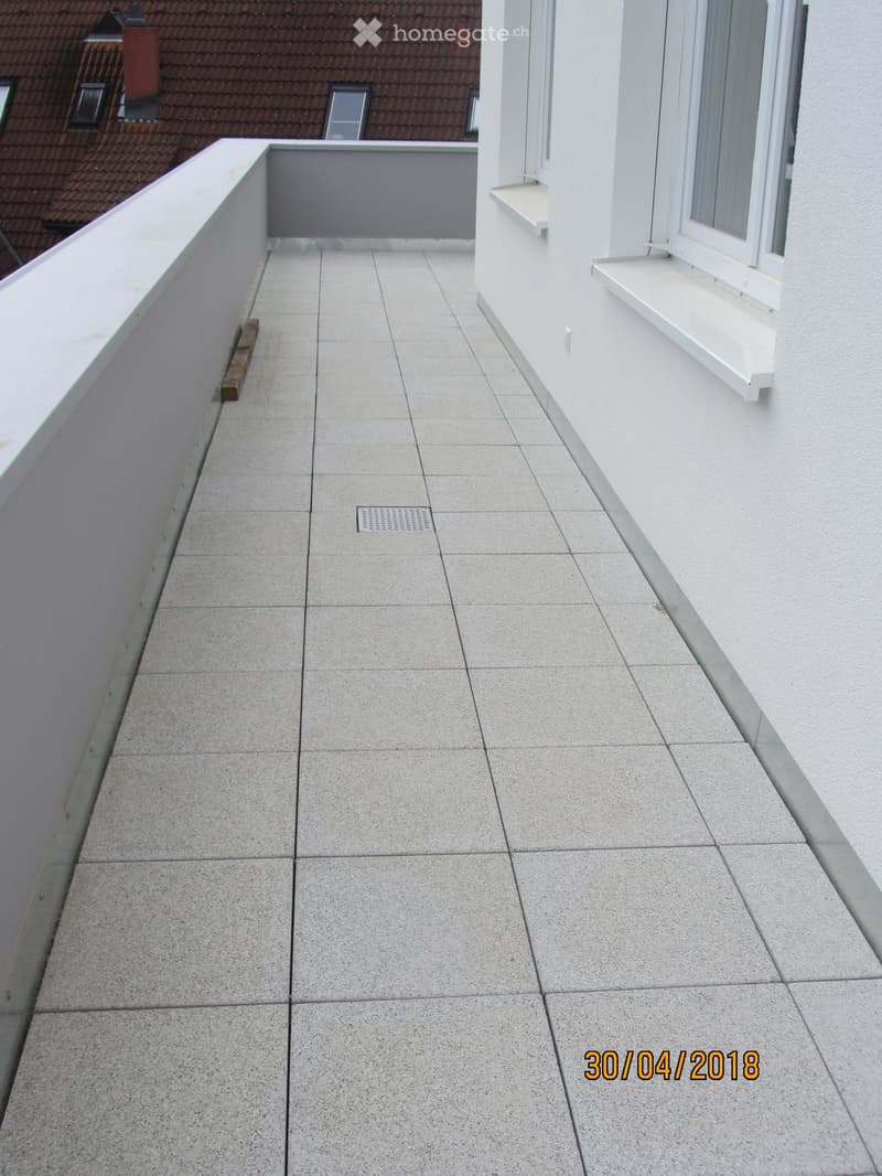Balcony with tiled floor, white walls, and a small window with a white sill. The balcony has a low railing and a small wooden block on the left side.