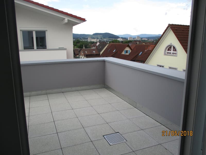 Empty balcony with tiled floor, low white railing, and a view of surrounding rooftops and distant hills.