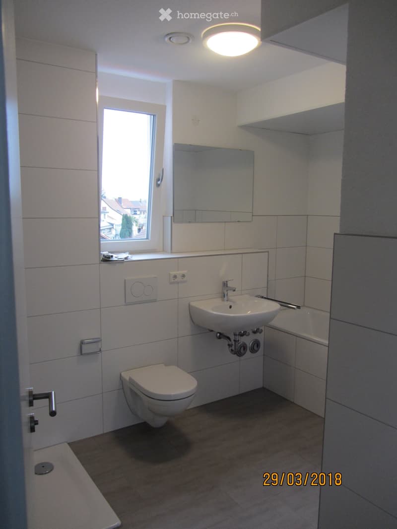 Modern bathroom with white tiles, a toilet, a sink with a mirror, and a window providing natural light.