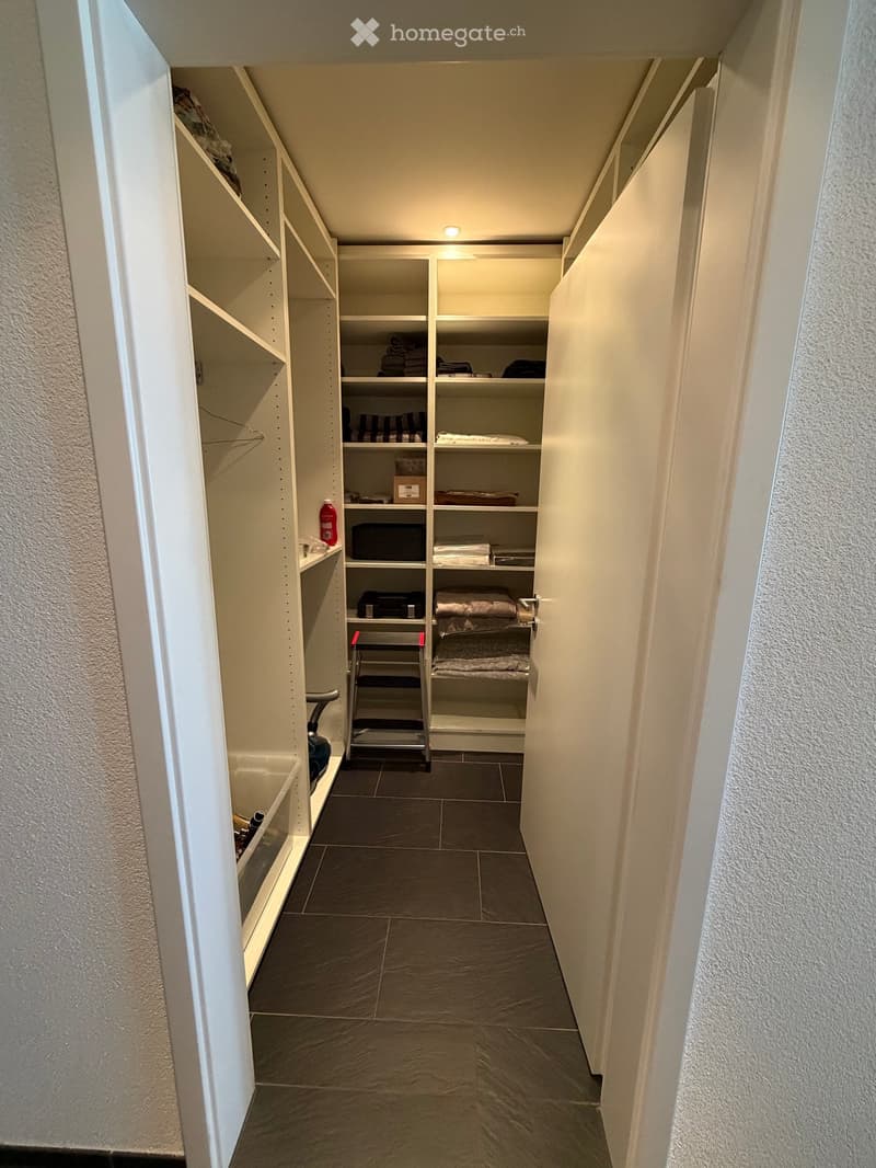 A narrow hallway with built-in white shelving units on both sides, containing folded towels, baskets, and other stored items. The floor is tiled in dark gray, and the walls are painted white.