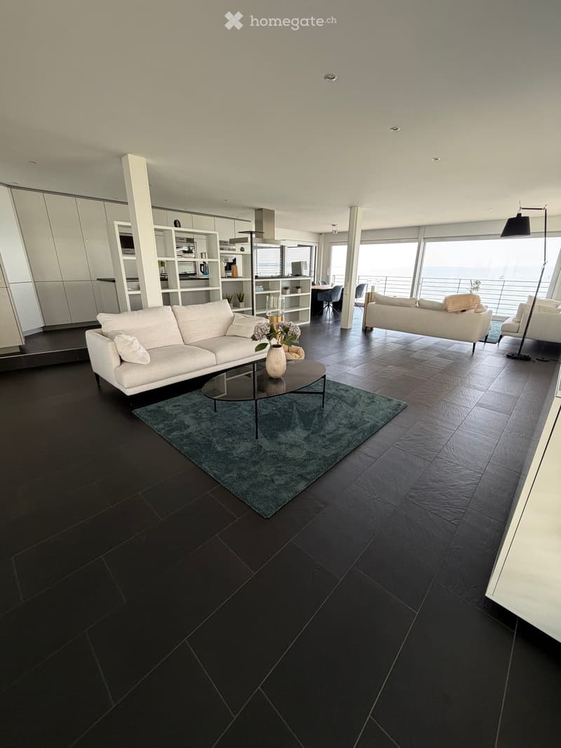 Modern living room with dark tiled flooring, white sofa, coffee table, vase with flowers, and a view of the ocean through large windows.