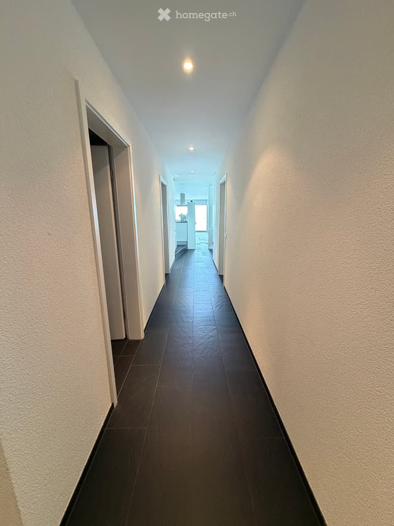 A long, narrow hallway with dark tiled flooring and light-colored walls, featuring several doors and recessed lighting.