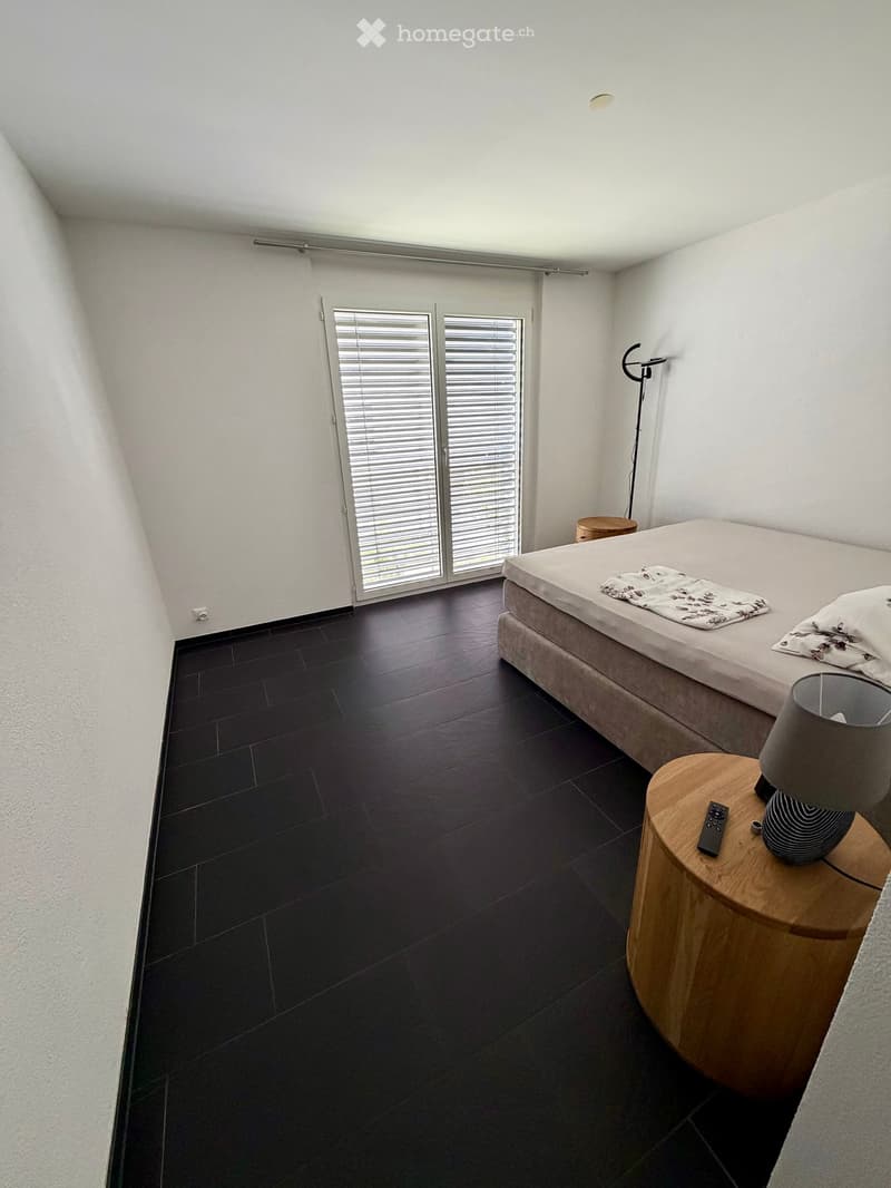 Modern bedroom with dark tiled floor, white walls, large window with blinds, bed with pillows and blanket, nightstand with lamp and remote control.