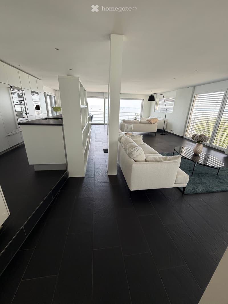 Modern living room with black tiled flooring, white walls, and large windows. Features a white sofa, coffee table, vase, floor lamp, and a kitchen island with cabinets.