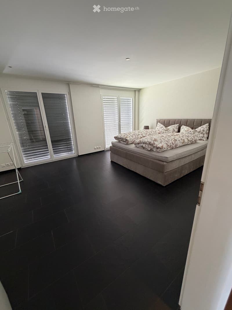 Modern bedroom with dark wooden flooring, large windows with blinds, and a neatly made bed with patterned bedding.