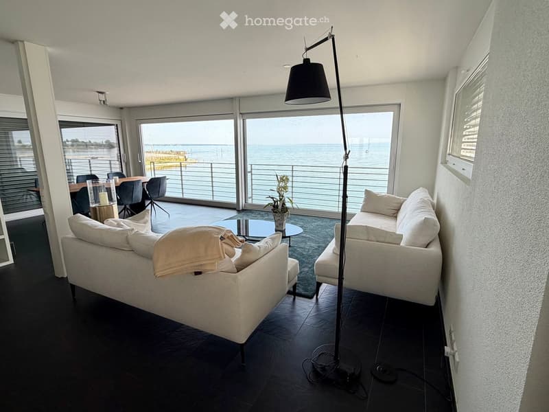 Modern living room with large windows offering a sea view, featuring a white sofa, coffee table, chairs, and a floor lamp.