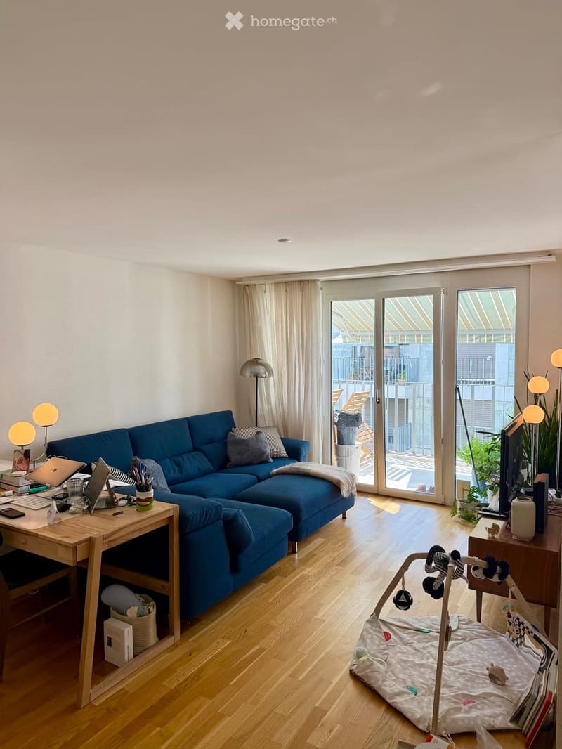 Modern living room with a blue sectional sofa, wooden floor, large sliding glass doors leading to a balcony, and a small desk with a laptop.