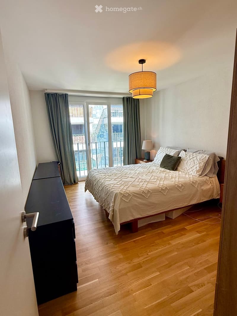 Modern bedroom with wooden flooring, large bed, nightstand, pendant light, and balcony access through large glass doors.