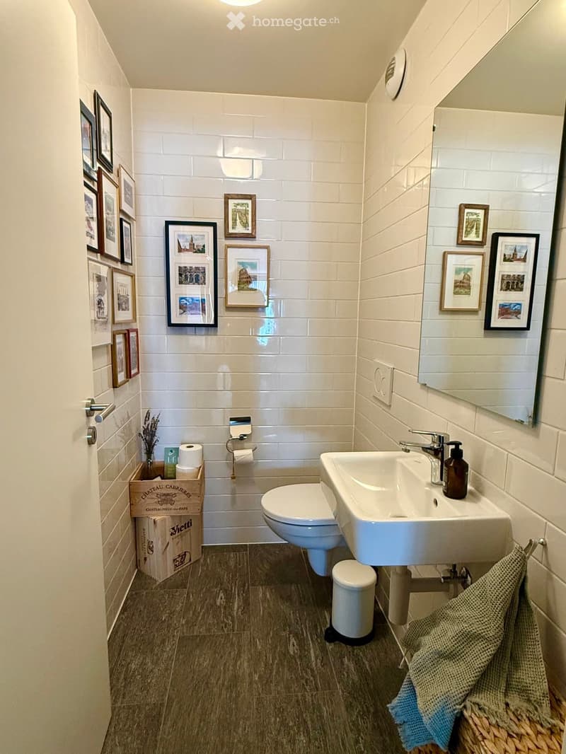 Modern bathroom with white tiled walls, a toilet, a sink with a modern faucet, a mirror, and framed pictures on the walls. A woven basket holds a towel, and a small wooden crate contains toilet paper and other items.