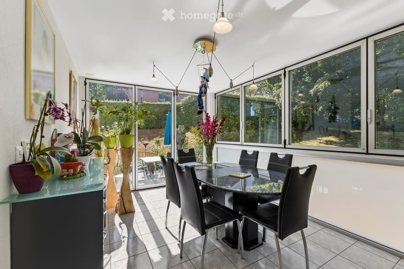 Bright dining area with a glass table, black chairs, potted plants, and large windows overlooking a garden.