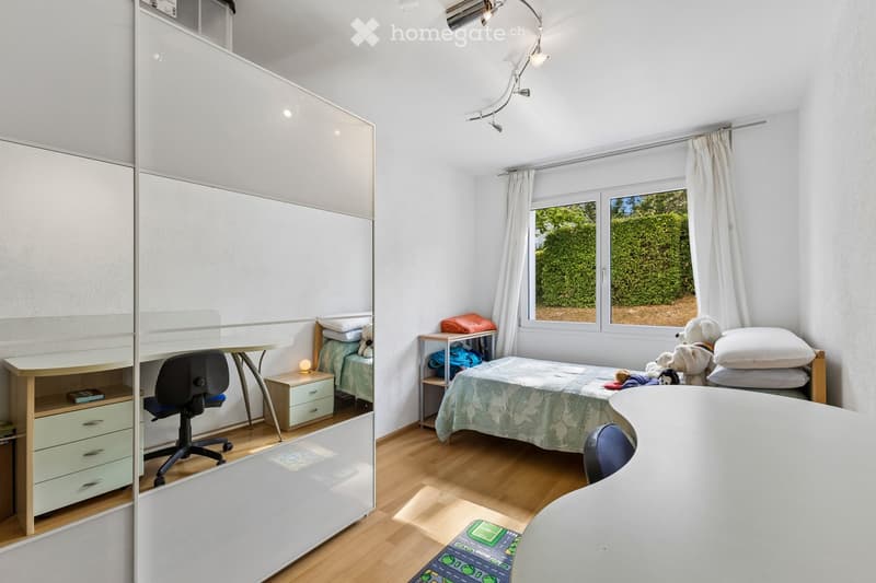 A well-lit bedroom with a bed, desk, chair, wardrobe, and nightstand. The room features wooden flooring and large windows with white curtains, allowing natural light to fill the space.