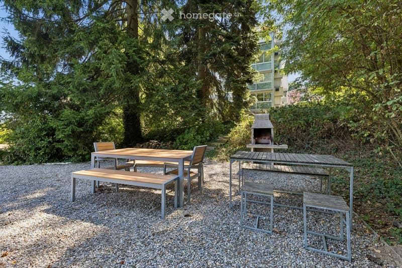 Outdoor seating area with a wooden table, benches, and chairs, surrounded by gravel and trees, with a barbecue in the background.