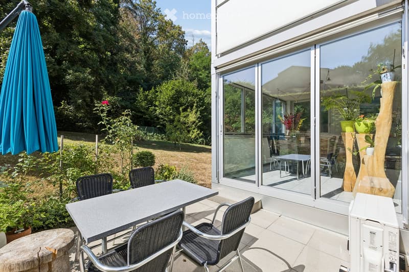 Balcony with a table, chairs, umbrella, and potted plants, adjacent to a modern house with large glass windows.