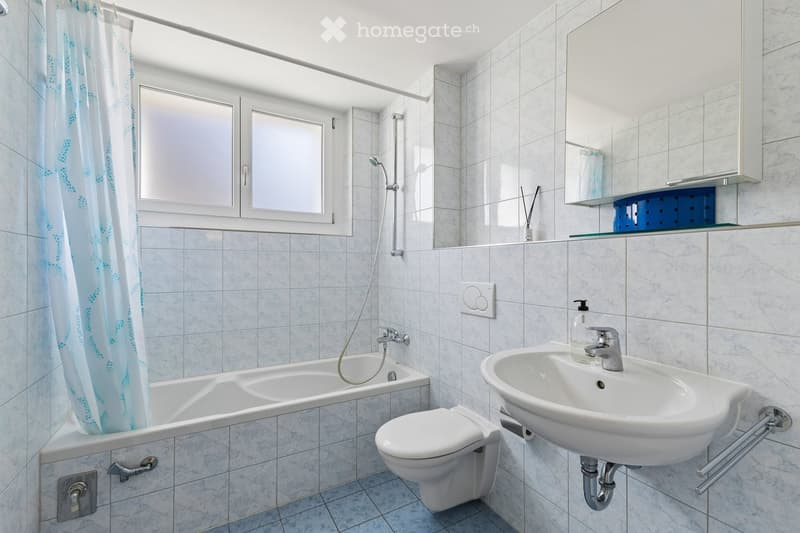 Modern bathroom with white tiles, a bathtub, toilet, sink, mirror, and a shower curtain. Features a window with frosted glass panes and a small shelf with decorative items.