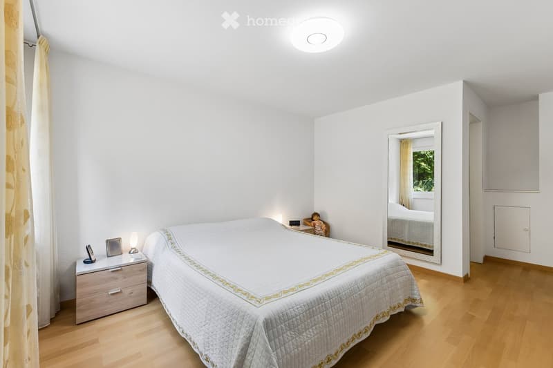 A modern bedroom with a neatly made bed, a nightstand with a lamp, a mirror, and a small table with a chair.