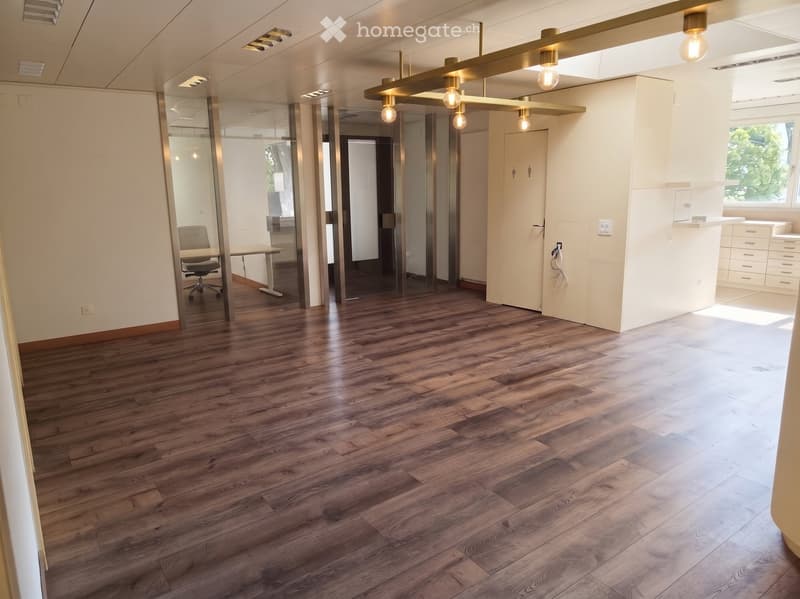 Open-plan office space with wooden flooring, glass partitions, a chair, a table, and a cabinet.
