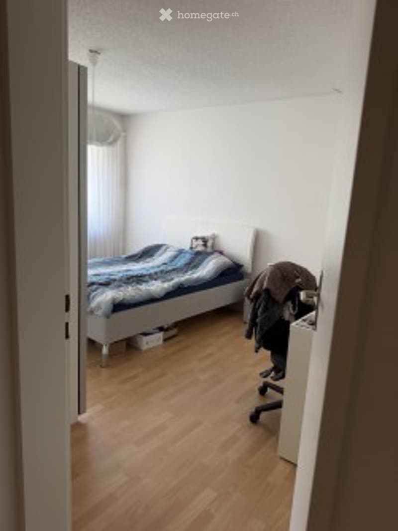 Minimalist bedroom with light wooden flooring, white walls, a single bed with a blue and white duvet, a small chair, and a wardrobe with hanging clothes.