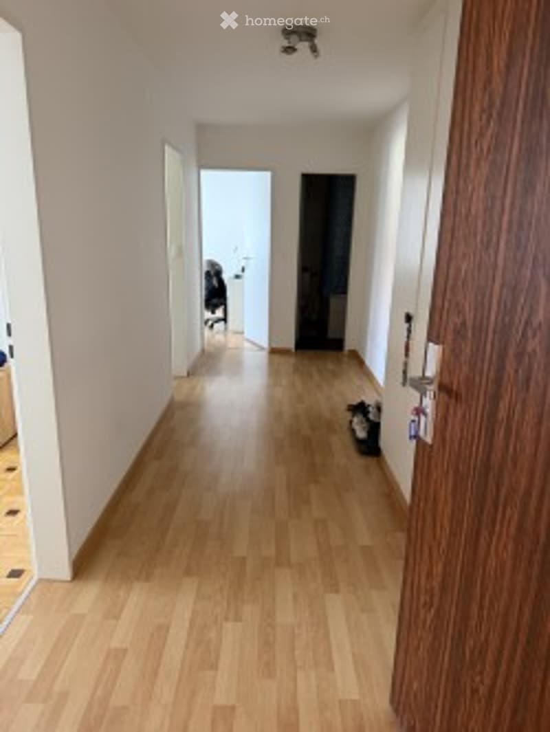 A long, narrow hallway with light wooden flooring and white walls. The hallway features a closed door at the far end and an open doorway leading to another room. A small black bag is placed near the open doorway.