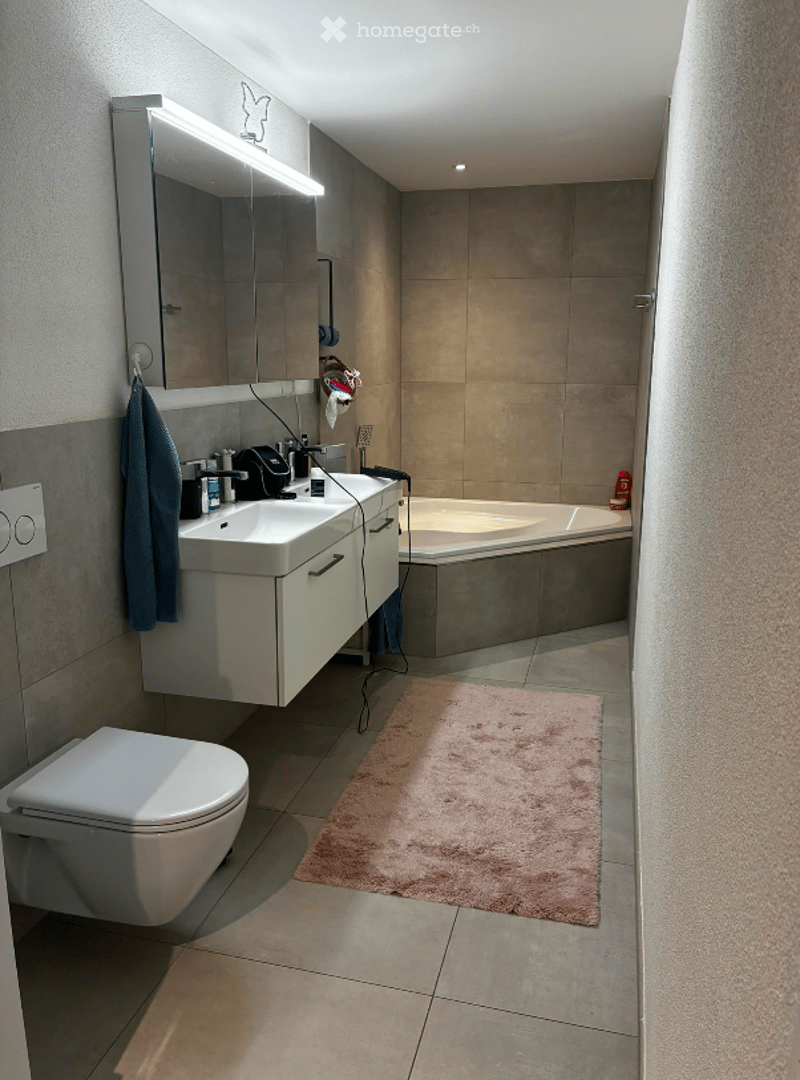 Modern bathroom with a white sink, mirror with LED lighting, bathtub, toilet, and a pink bathroom rug on the floor.