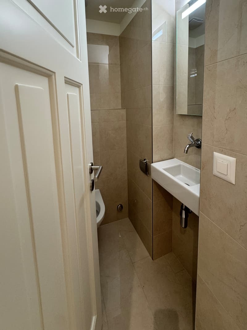 Modern bathroom with beige tiles, a white sink, a chrome faucet, a mirror, and a toilet. The space is compact with a small window at the top.