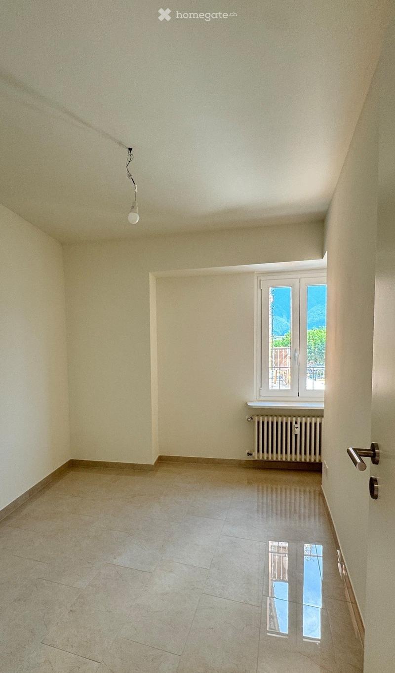 Empty room with light-colored walls, tiled floor, and a window with a radiator below it. A single light fixture hangs from the ceiling.