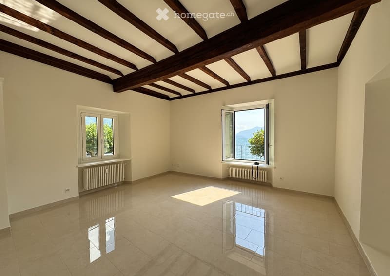 Empty room with exposed wooden beams on the ceiling, large windows offering a scenic view, tiled floor, and radiators under the windows.