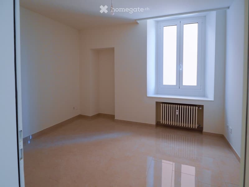 Empty room with tiled floor, white walls, large window, radiator, and a small built-in shelf.