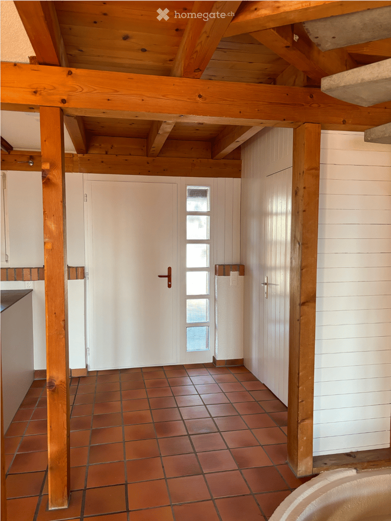 Bright hallway with wooden beams and exposed ceiling, red tiled floor, white walls, and a door with glass panels.