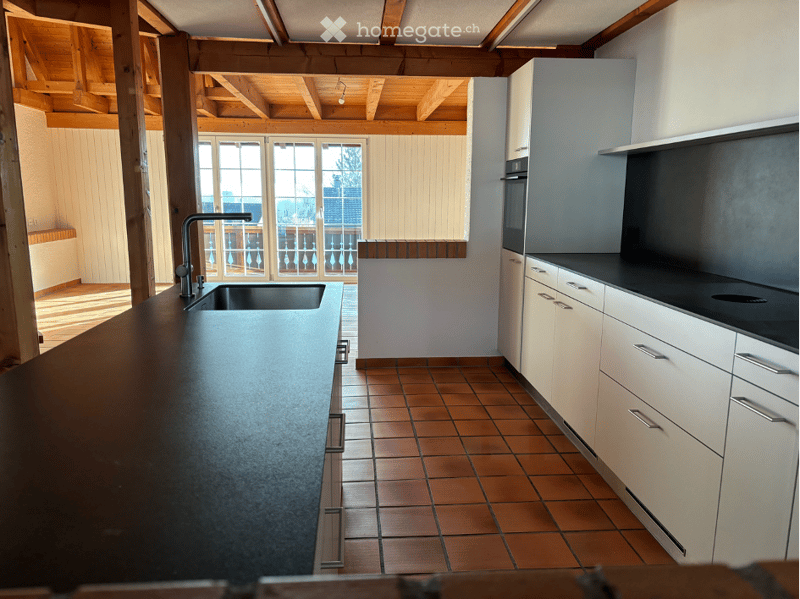 Modern kitchen with white cabinets, dark countertop, stainless steel sink, and built-in oven. Features exposed wooden beams on the ceiling and a large window with a view to a balcony.