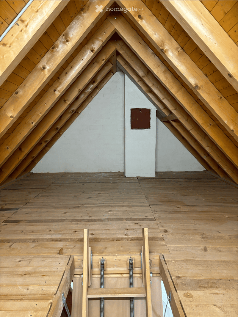 An attic space with exposed wooden beams and a wooden floor, featuring a small access door and visible insulation panels.