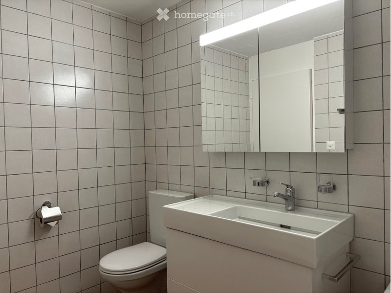 Modern bathroom with white tiled walls, a white sink with a chrome faucet, a large mirrored cabinet with built-in lighting, and a toilet. The space is clean and contemporary with a minimalist design.