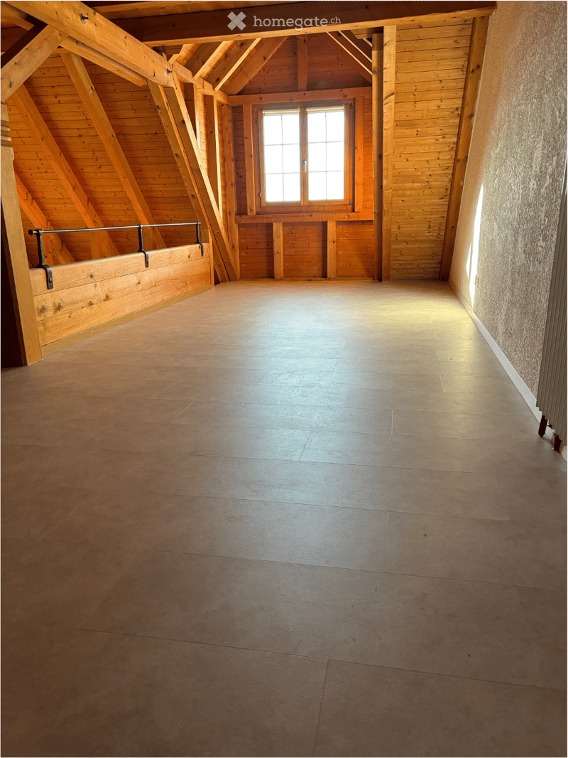Empty attic space with wooden beams, exposed wooden ceiling, and a single window letting in natural light. The floor is made of polished concrete tiles.