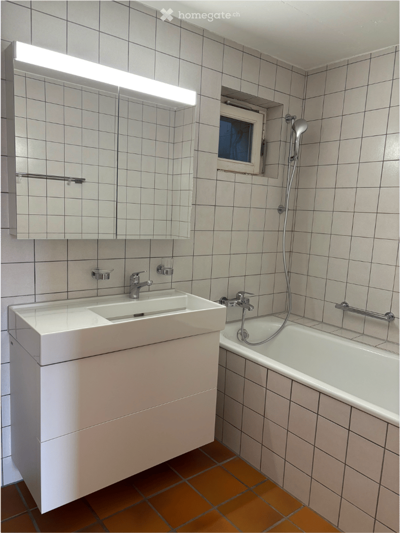 Modern bathroom with white tiled walls, a white sink with a chrome faucet, a bathtub with a handheld showerhead, and a large mirror with built-in lighting above the sink.