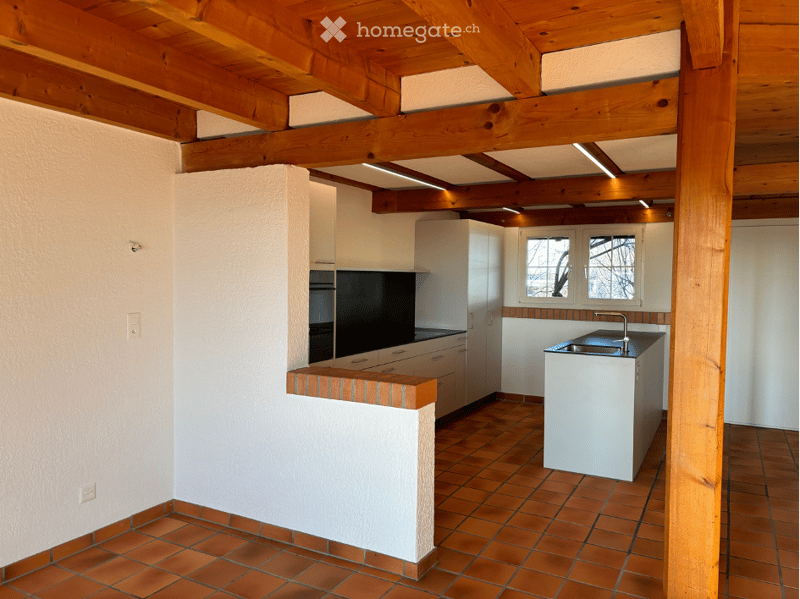 Modern kitchen with white cabinets, a central island, and a sink. The space features exposed wooden beams on the ceiling and terracotta tiled flooring.